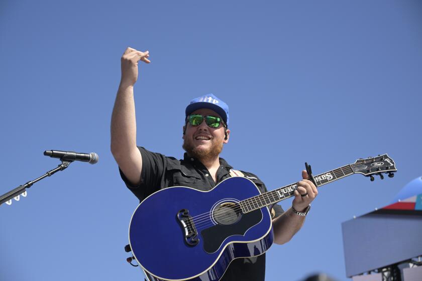 A man in a blue hat and sunglasses holding a blue acoustic guitar and raising his right hand in front of a microphone