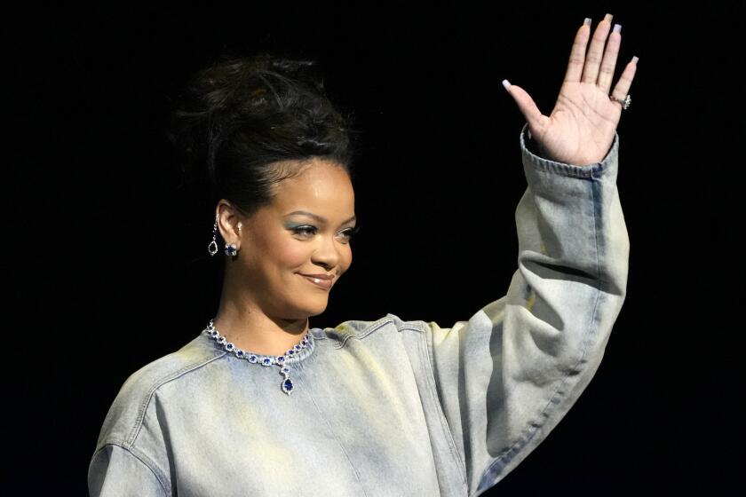 Rihanna in a long jean dress holding up her left hand while standing on a stage
