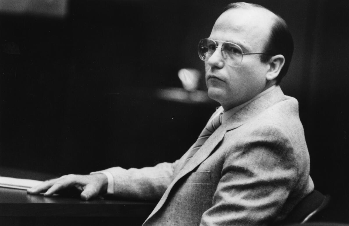 A black-and-white photo of a balding man in aviator-frame glasses and a suit, looking unhappy