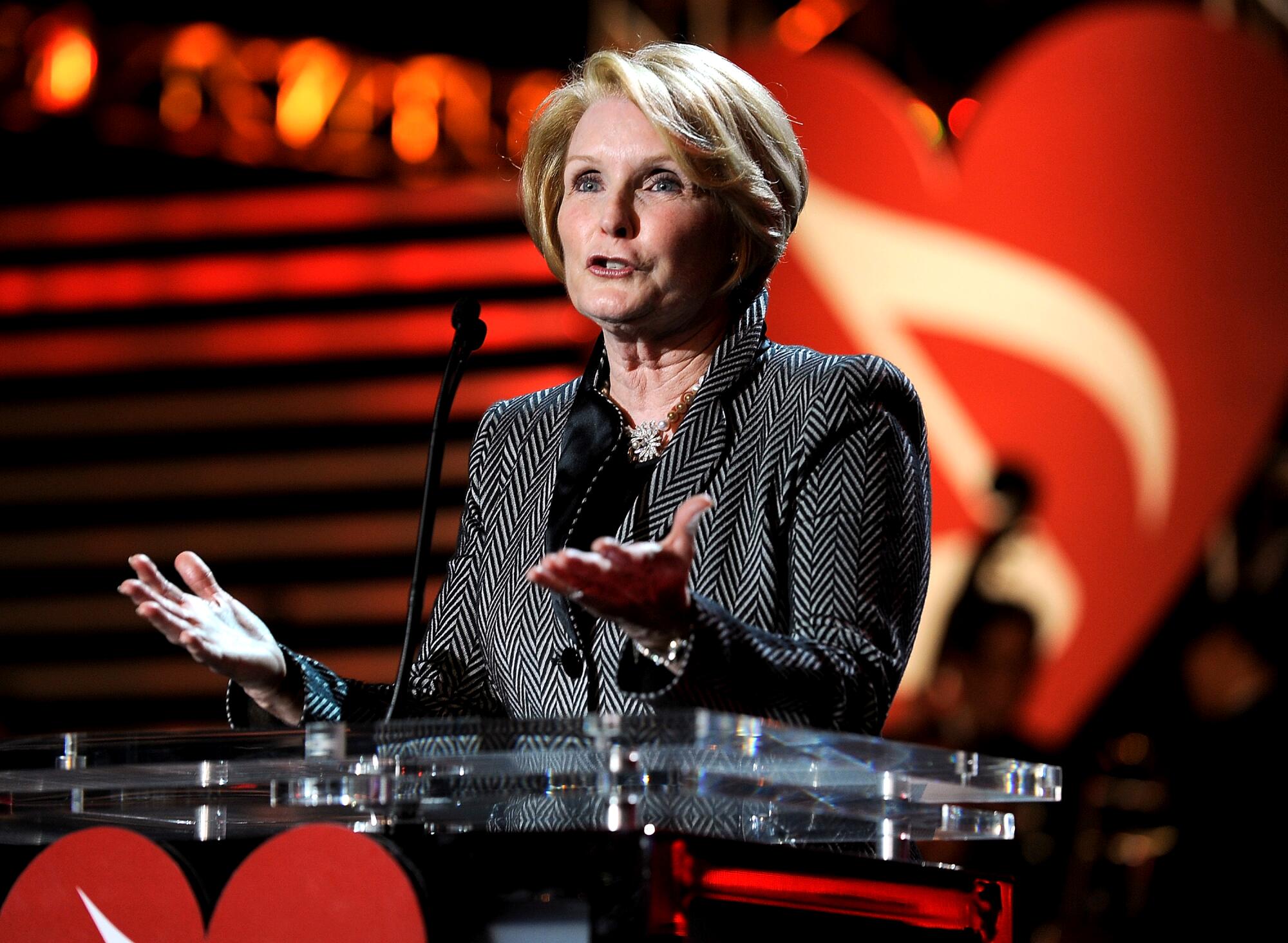 A blond woman stands at a transparent podium gesturing with both hands as she speaks.