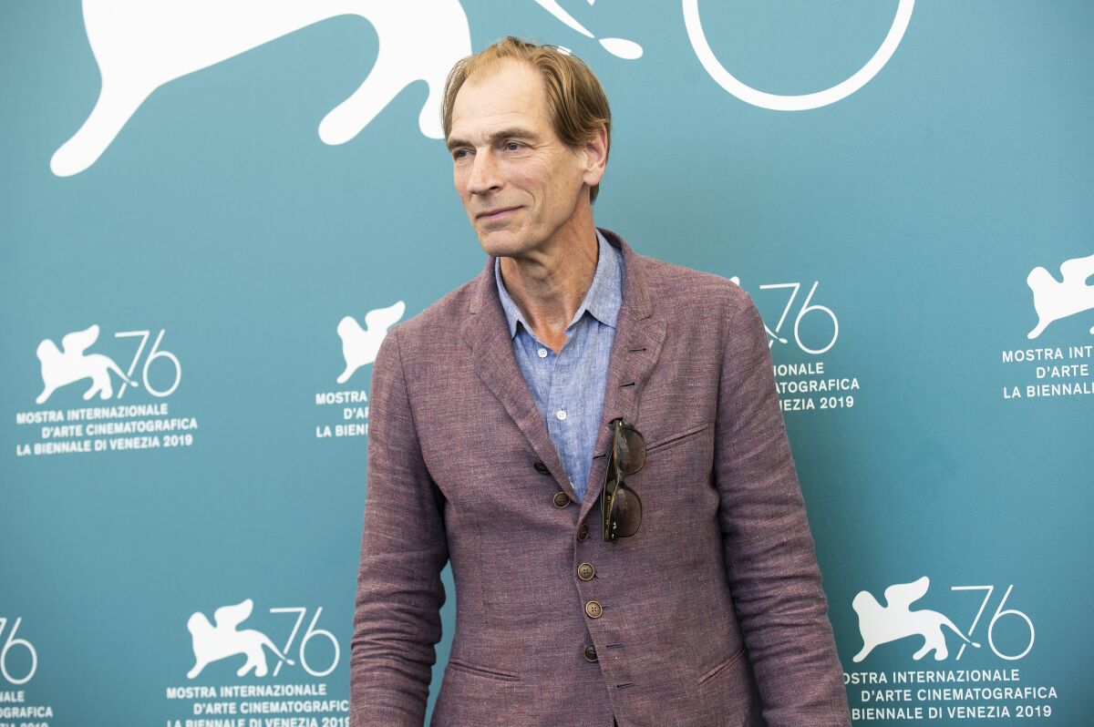 A man in a purplish blazer and blue shirt stands against a turquoise backdrop
