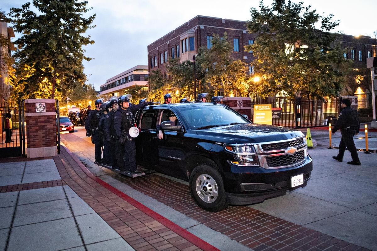A group of police officers wearing riot gear stands on the sides of an SUV that is traveling on the USC campus.