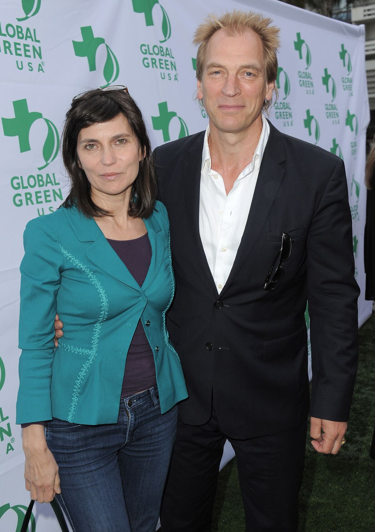 A woman in a green jacket and jeans poses with a  man in a white button-down shirt and black suit.