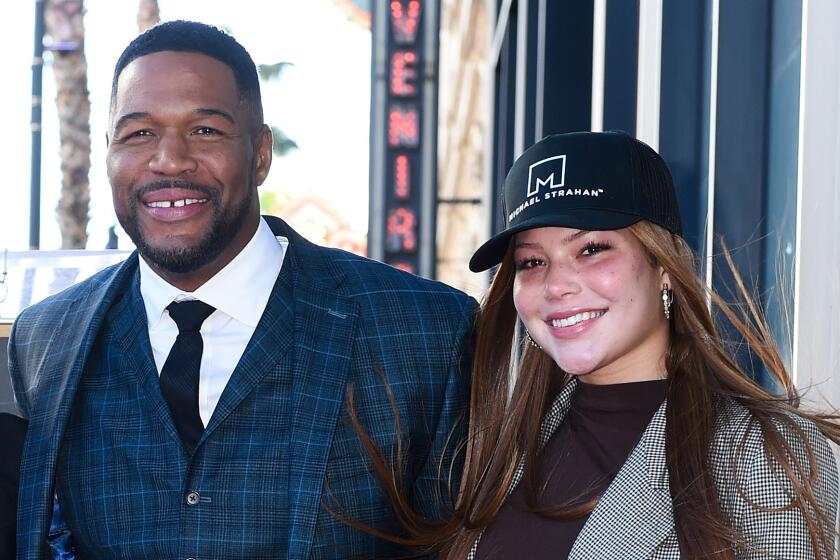 Michael Strahan in a blue plaid suit smiling next to a young woman with long hair and a black hat in a grey blazer