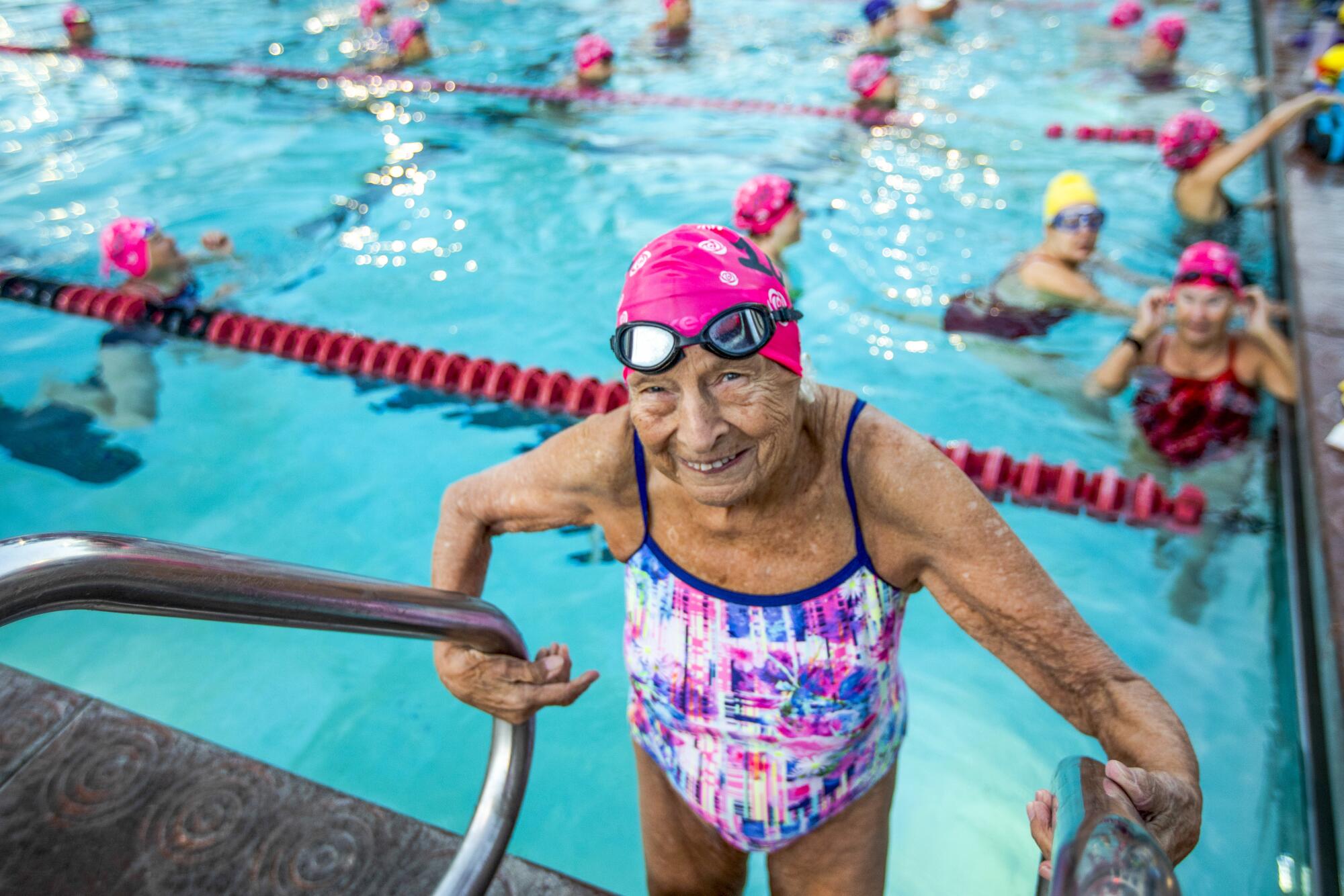 A 100-year-old woman in a multicolored bathing suit stands at the edge of a pool where other swimmers are in the water