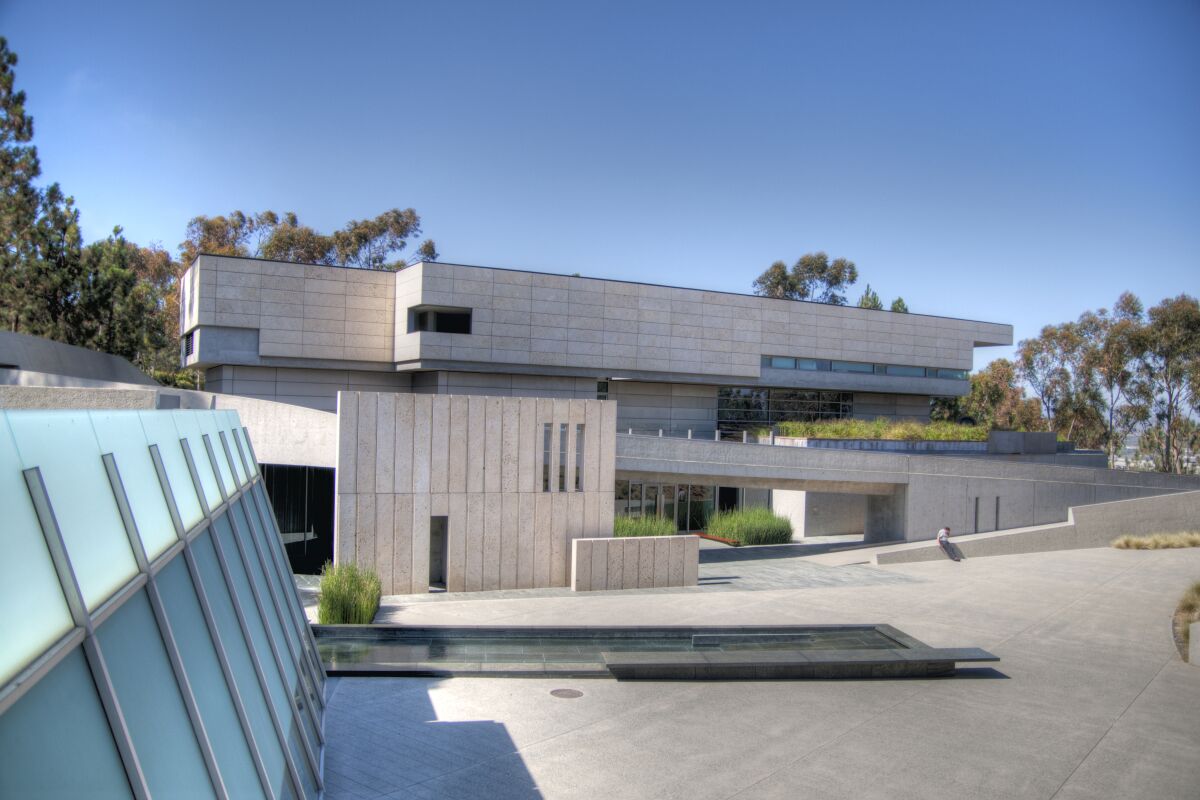 A horizontal building with a rectangular geometric facade made of sand-colored concrete emerges from a sloped hillside