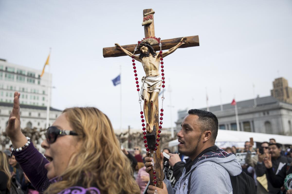 A man, right, holds up a crucifix with a red beaded necklace. Next to him, a woman waves. 