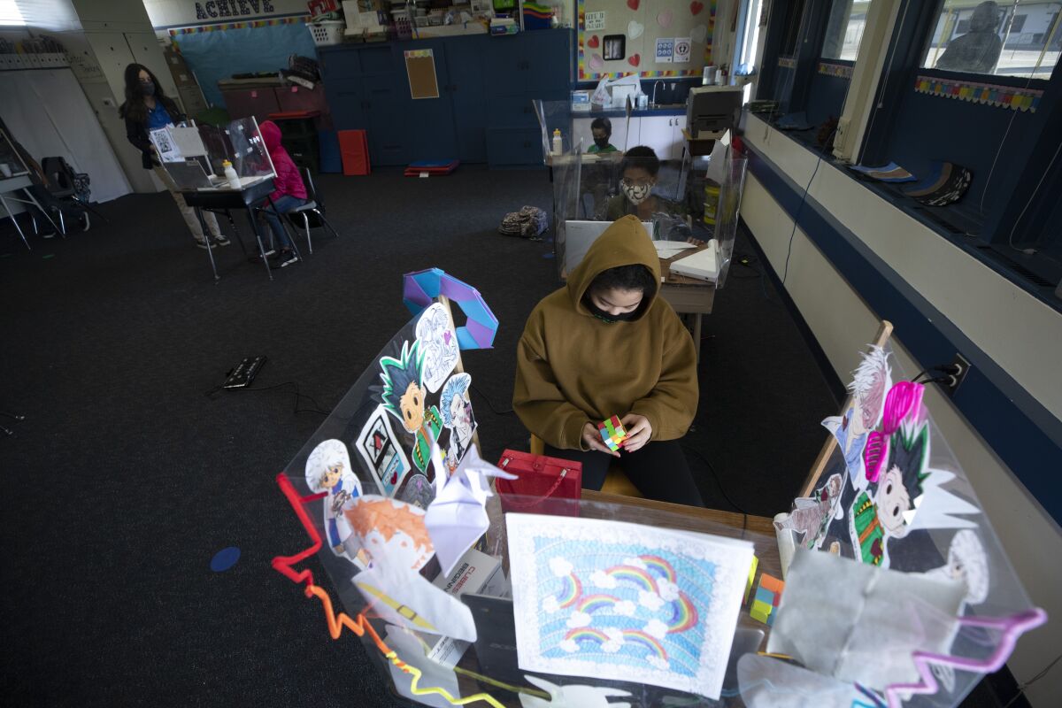 A girl in a mask and hoodie holds a Rubik's Cube at a desk with acrylic plastic dividers covered by drawings.