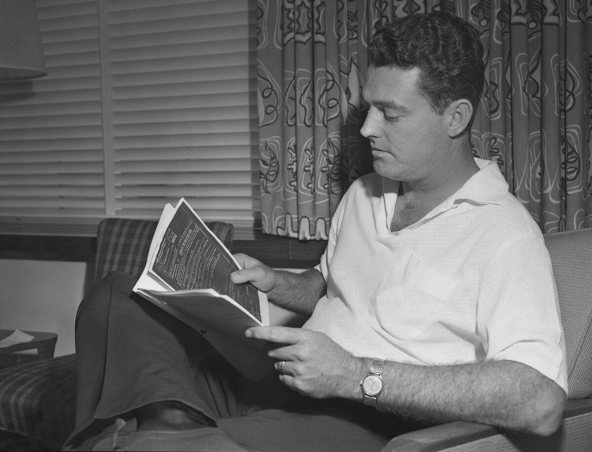 A man in a short-sleeve shirt sits in a chair with an ankle crossed over a knee, reading a packet of papers.
