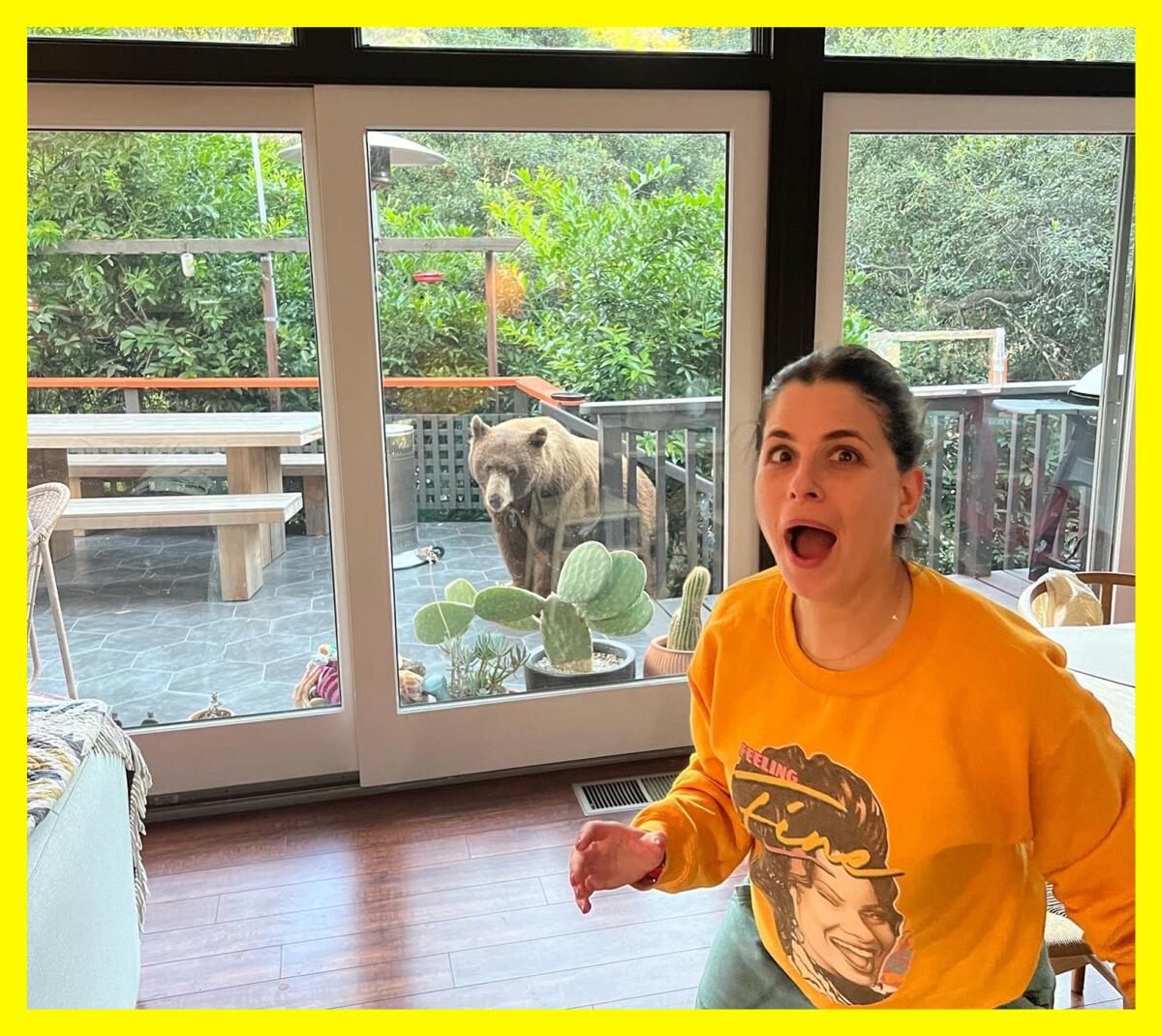 A woman in a living room, mouth agape, with a bear behind her outside a sliding glass door.