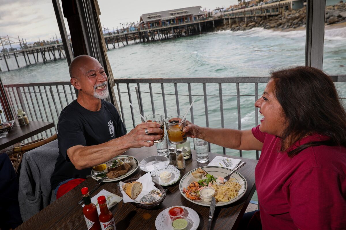A man and women clink their drink glasses over a table of food at a restaurant on the ocean