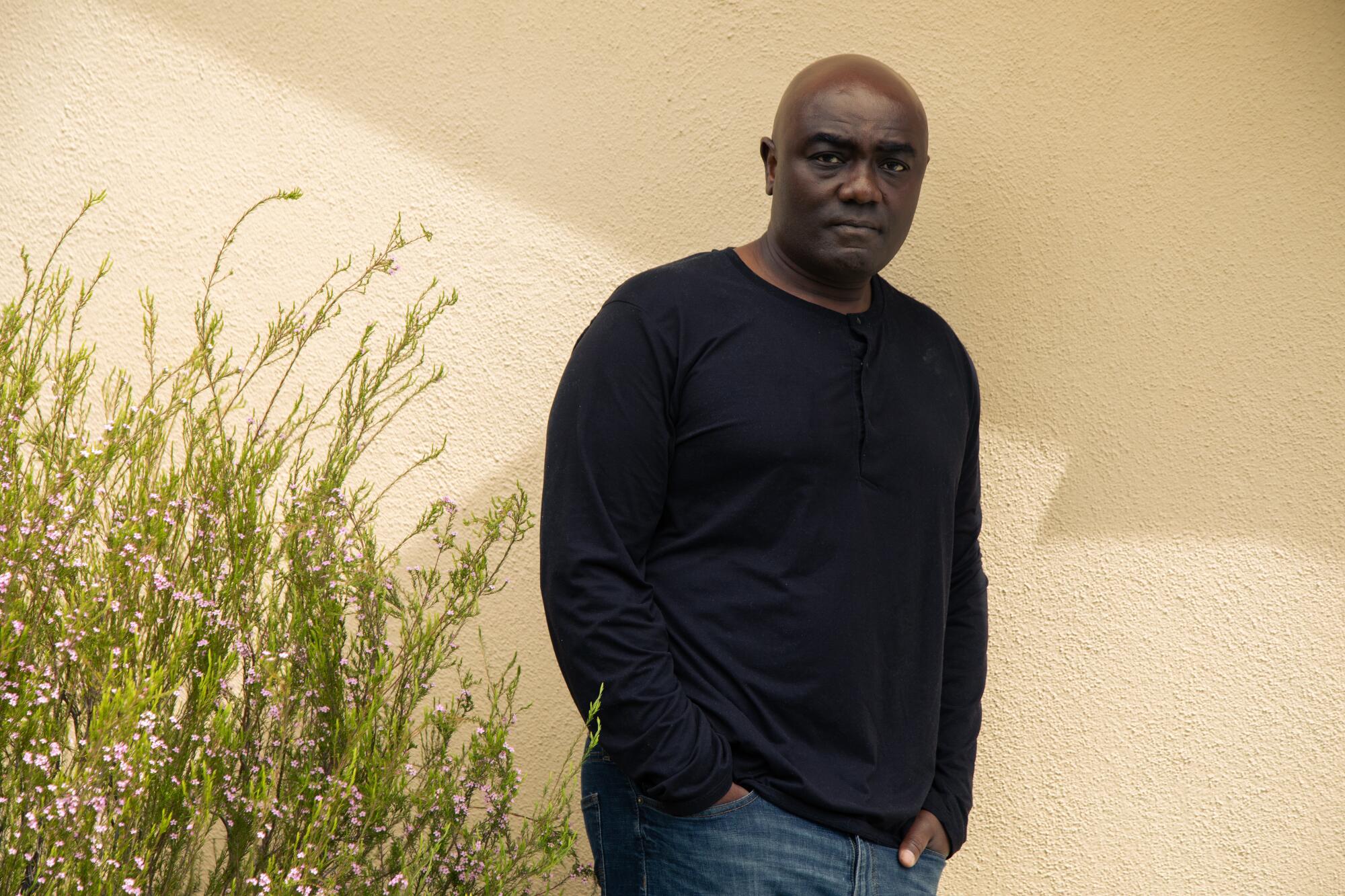 A man in a black sweatshirt and jeans stands against a yellow wall next to a flowering shrub.