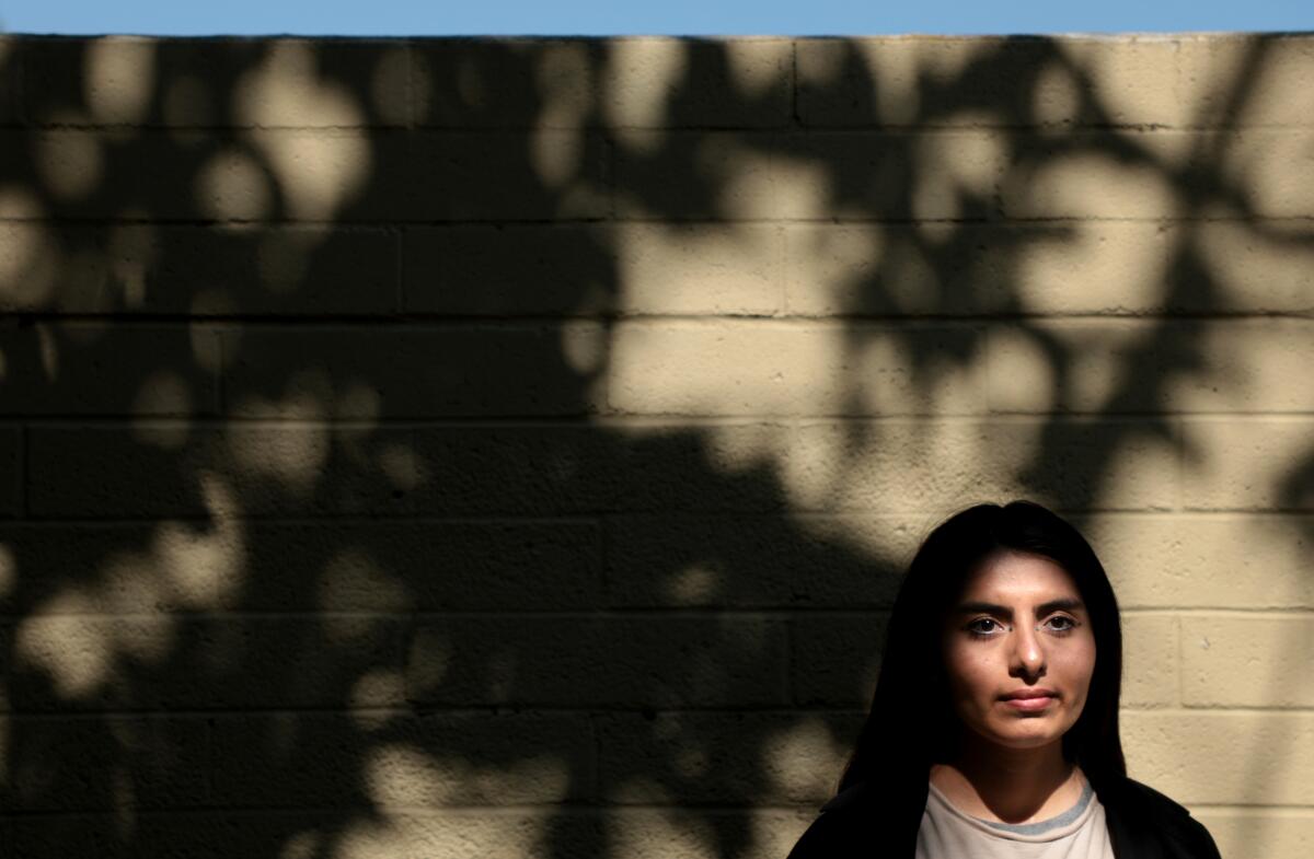 A young person with long black hair poses next to an empty brick wall.