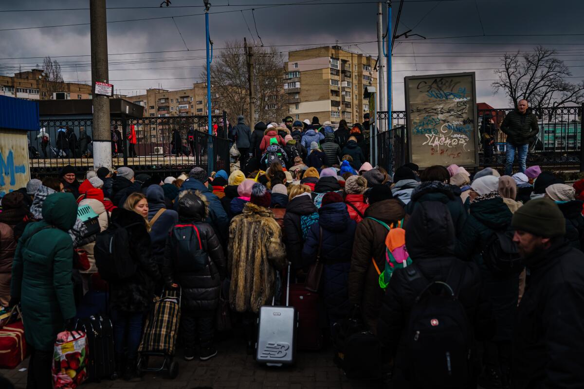 A crowd of people wearing jackets and hats approaching a train station gate. 