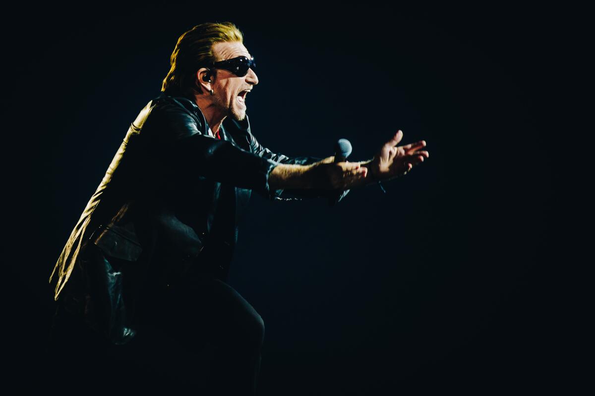 U2's Bono in black shades and jacket holding out his arms. 