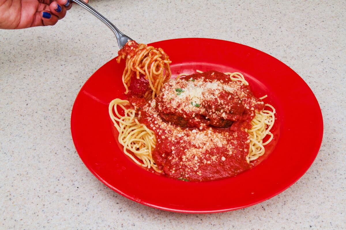 A hand pulls a twisted forkful of spaghetti from a red plate with meatballs and spaghetti.