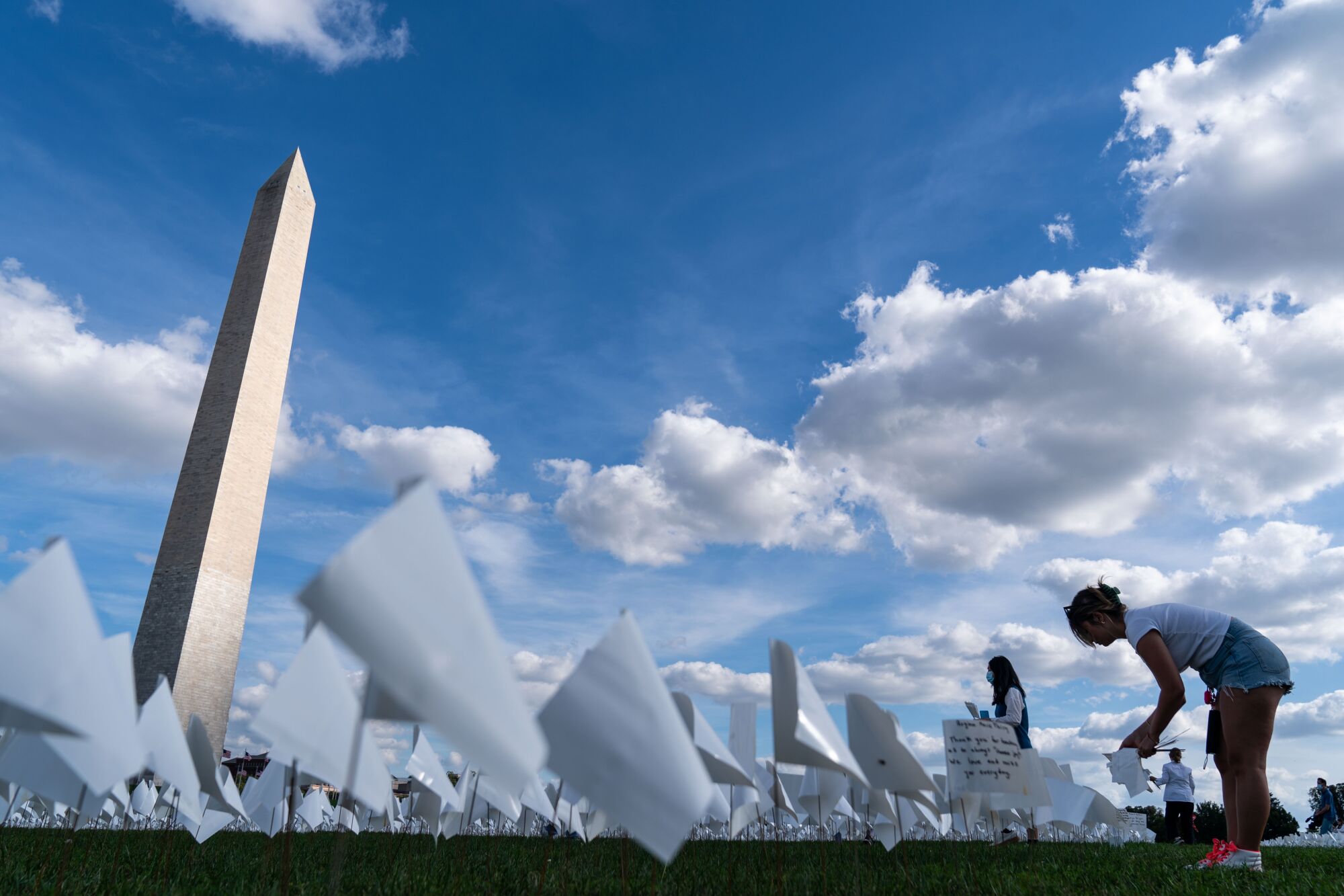 A woman bends over to place a small white flag in the grass, near the Washington Monument