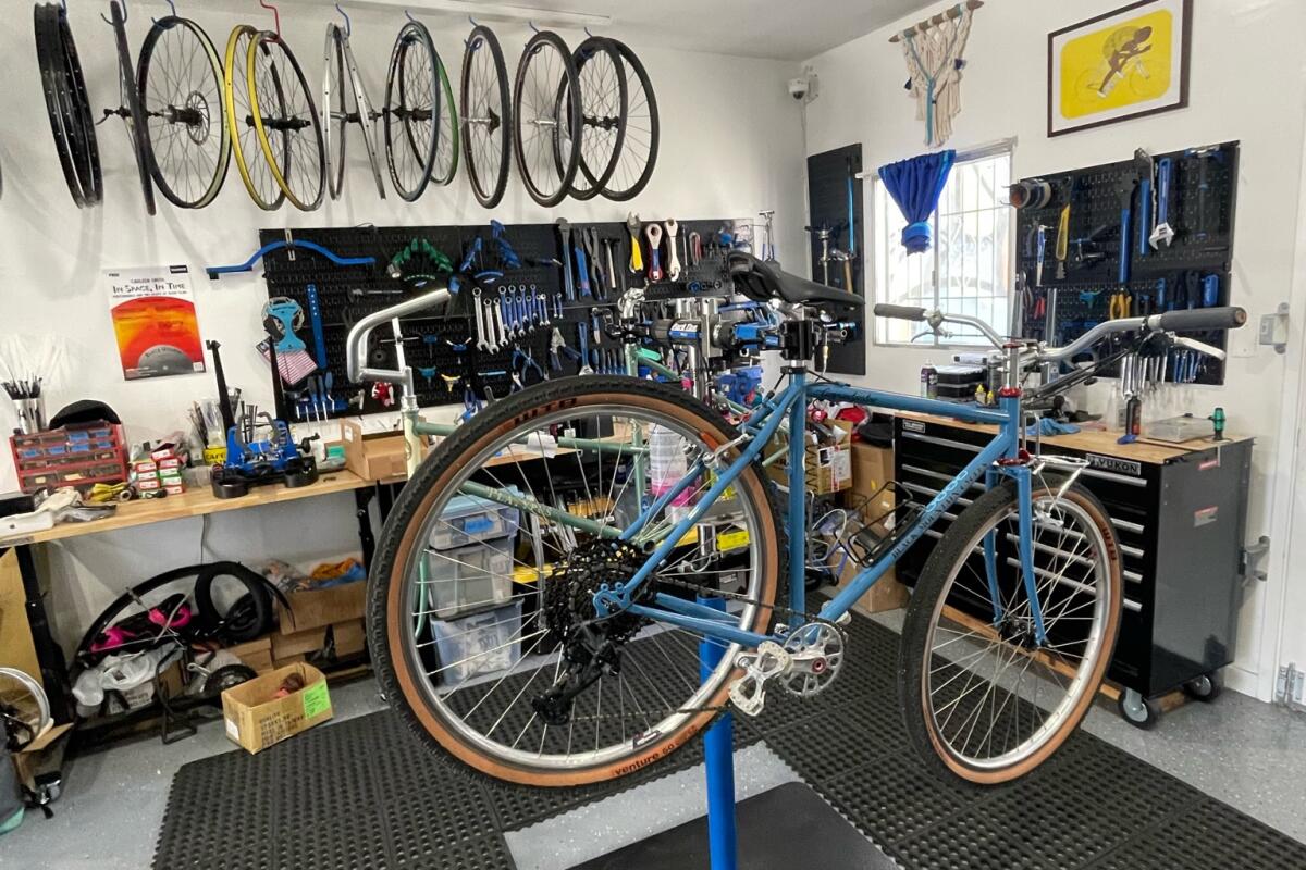 A bike on a stand in the center of a shop, with wheels hanging on the wall above.