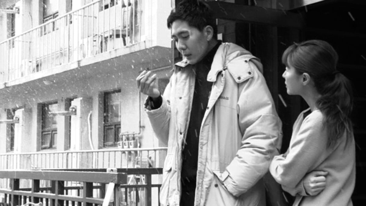 A man and a woman stand outside near an apartment building amid snow. He wears a jacket; she holds her arms in tight.
