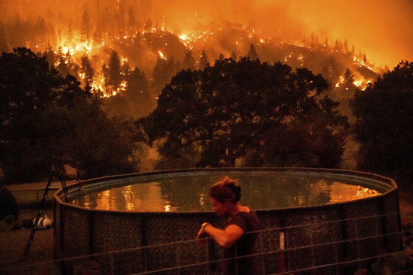 A woman leans against a fence next to an above-ground pool as wildfire burns a nearby ridge.
