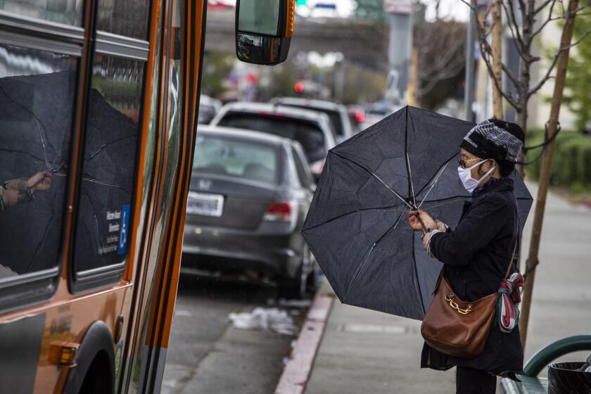 LOS ANGELES, CA - APRIL 18: A woman with an umbrella and a mask waits in the rain for a Metro bus at a stop on Hoover St. in the Pico Union neighborhood on Saturday, April 18, 2020 in Los Angeles, CA. Los Angeles County's busiest transit agency will make major cuts to bus and rail service starting Sunday. The cuts are the equivalent of a 29% annual reduction in bus service and a 14% reduction in rail service, officials said. (Brian van der Brug / Los Angeles Times)