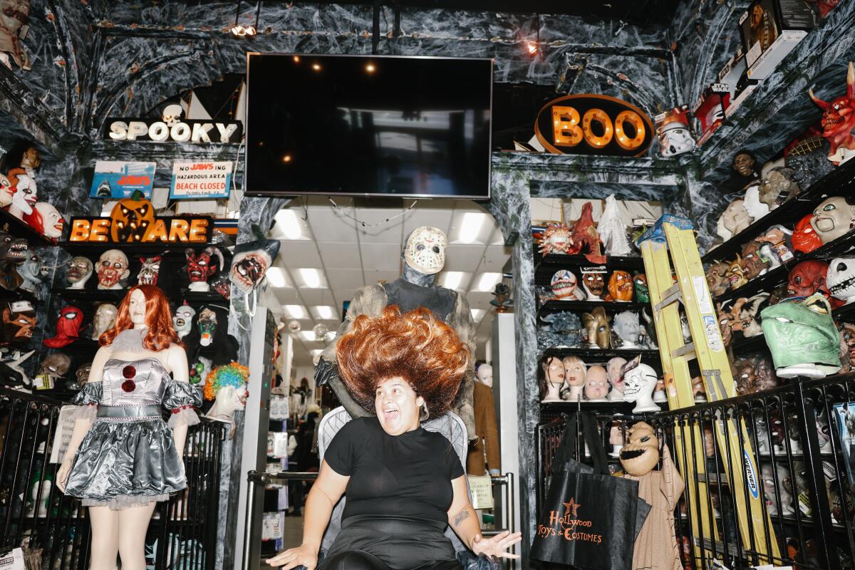 A woman pretends to scream while wearing a large wig in a room full of Halloween masks.
