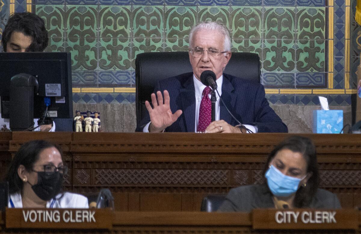 City Council President Paul Krekorian, wearing a black blazer and red tie, sits with his hand raised. 