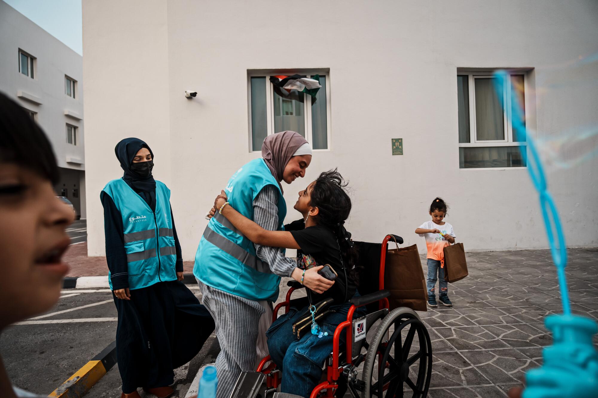 A child in a wheelchair hugs a young woman, in a hijab and turquoise vest, standing in a courtyard near other people