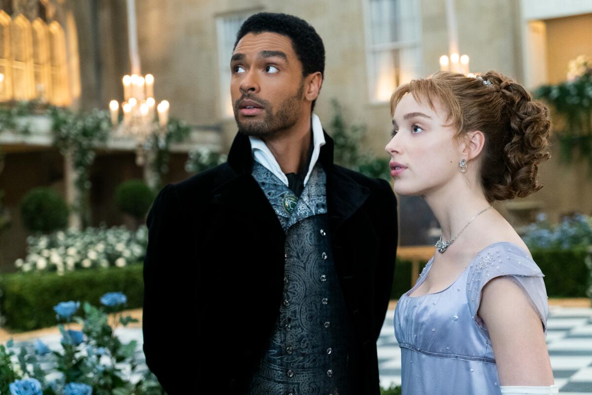 A man and a woman dressed in period evening clothes stand in a courtyard.