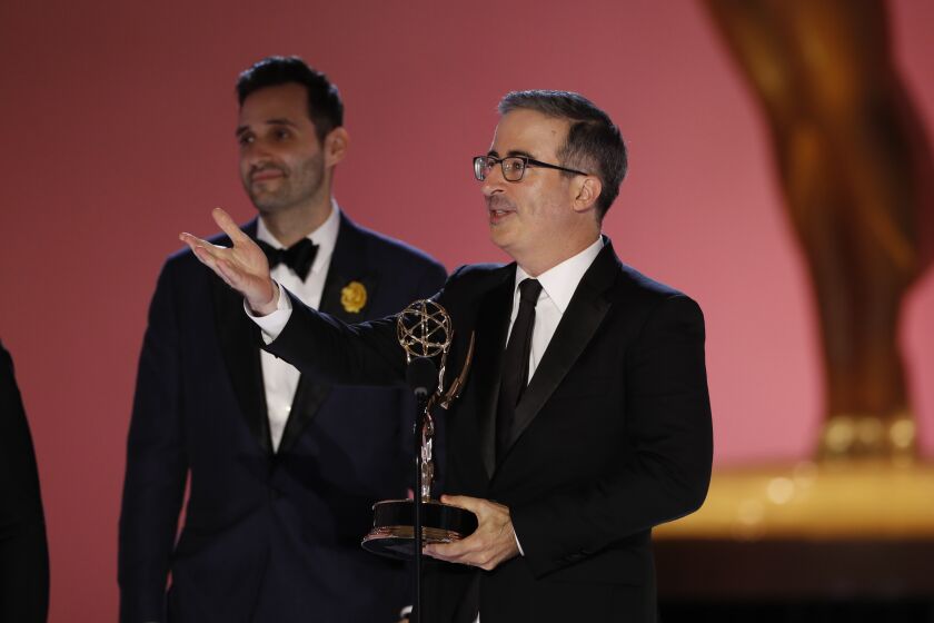 A man holding an Emmys trophy while speaking into a microphone and gesturing with his arm outstretched