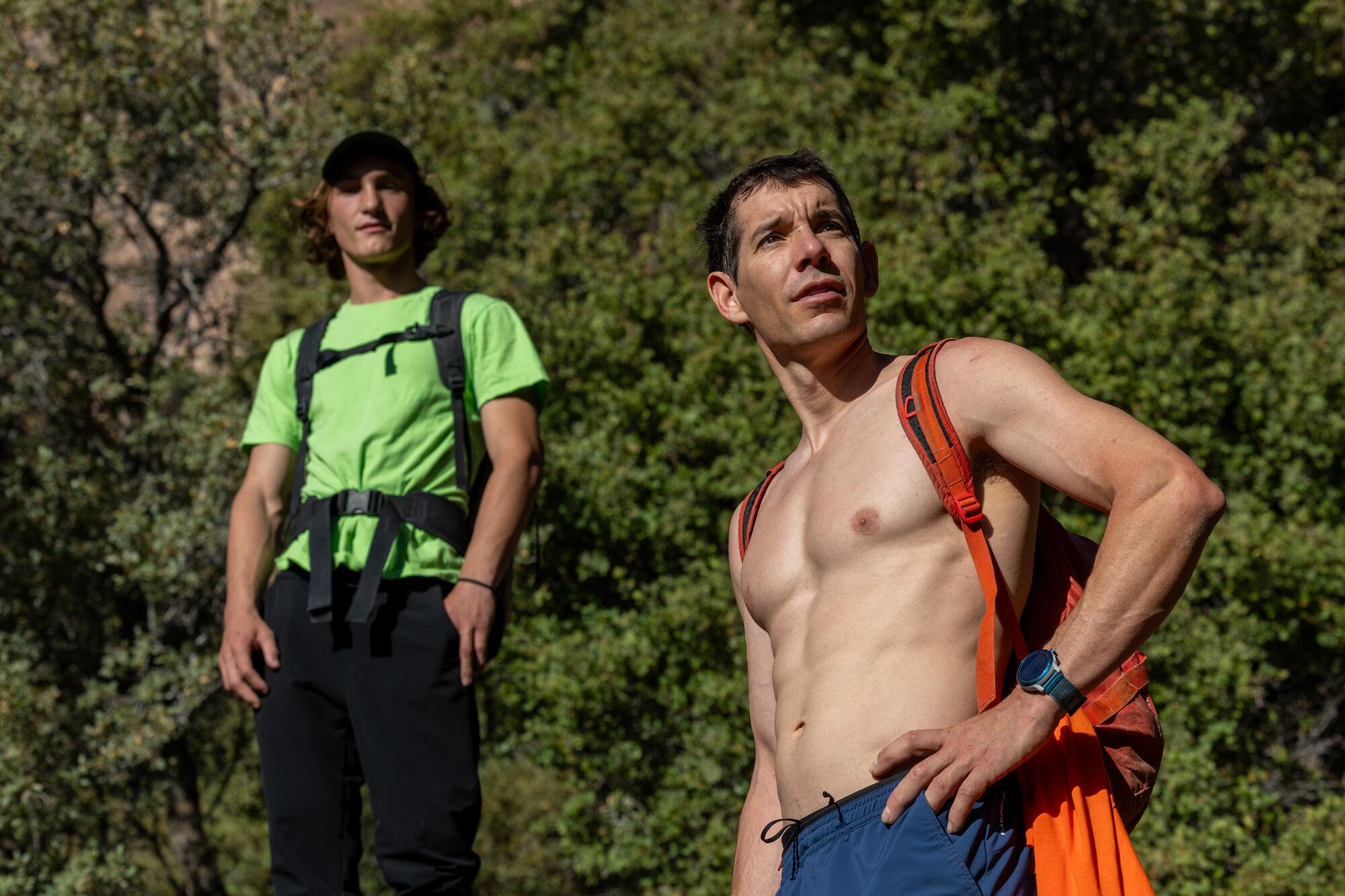 A fit, shirtless man and his hiking partner in a green T-shirt pause on a rugged canyon trail. 