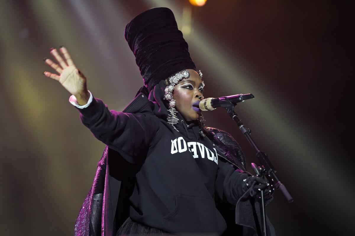 Lauryn Hill performs onstage wearing a large cylindrical black headpiece, purple cape and black sweatshirt