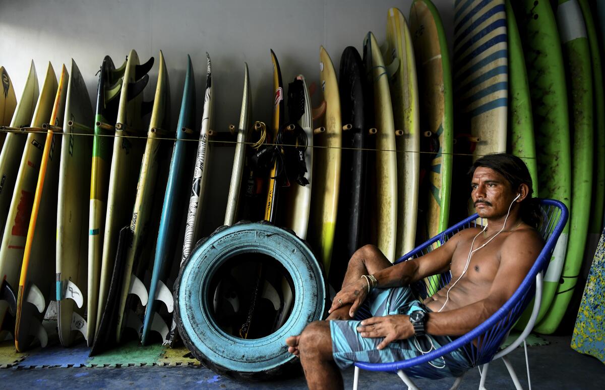 Edwin Omar Zepeda Morales, in swim trunks, sits before a rack of surfboards.