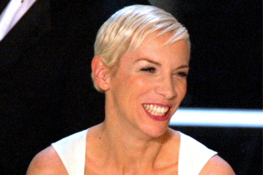 Singer Annie Lennox is wearing a white dress and smiling as she holds her Oscar