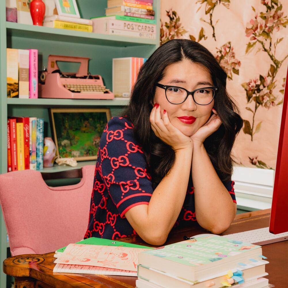 Jenny Han, in a navy blue and red Gucci dress, sits at a desk with her hands framing her face.