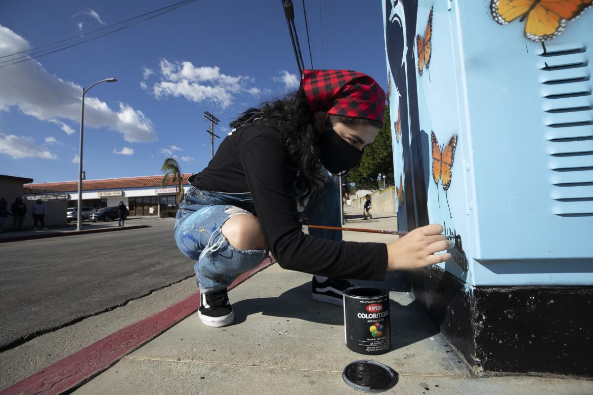 A young woman squats and uses a small brush to sign the bottom of her mural.