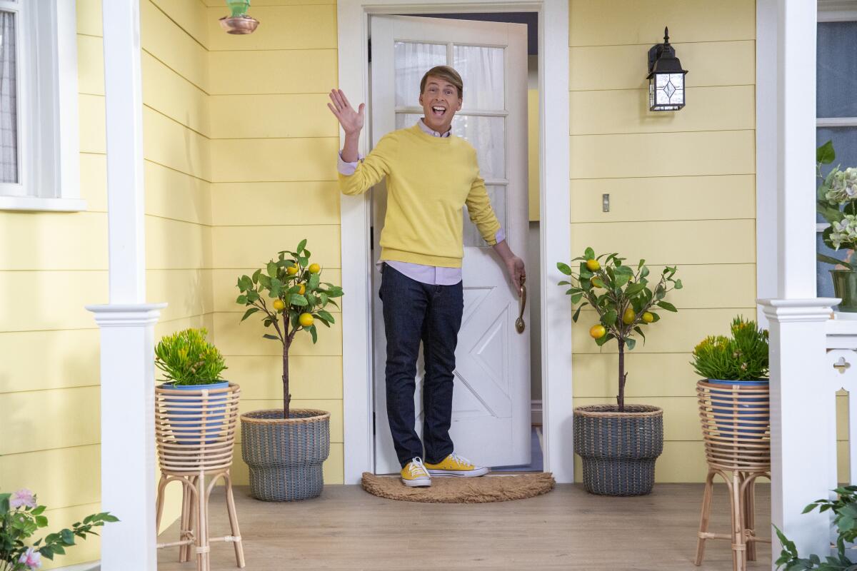 A man in a yellow sweater waves from the porch of a yellow house