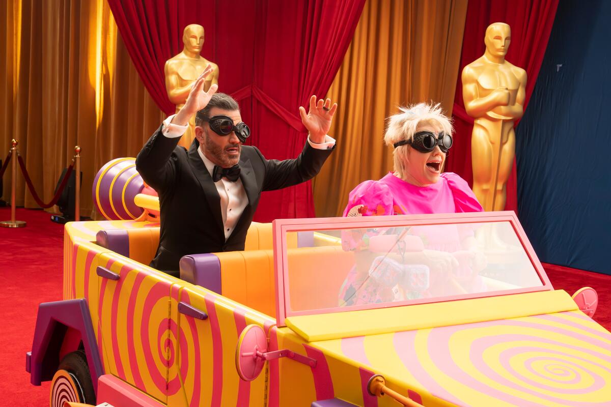 Jimmy Kimmel rides in the back of a jeep as Weird Barbie (Kate McKinnon) drives around the Oscar stage, both wearing goggles.