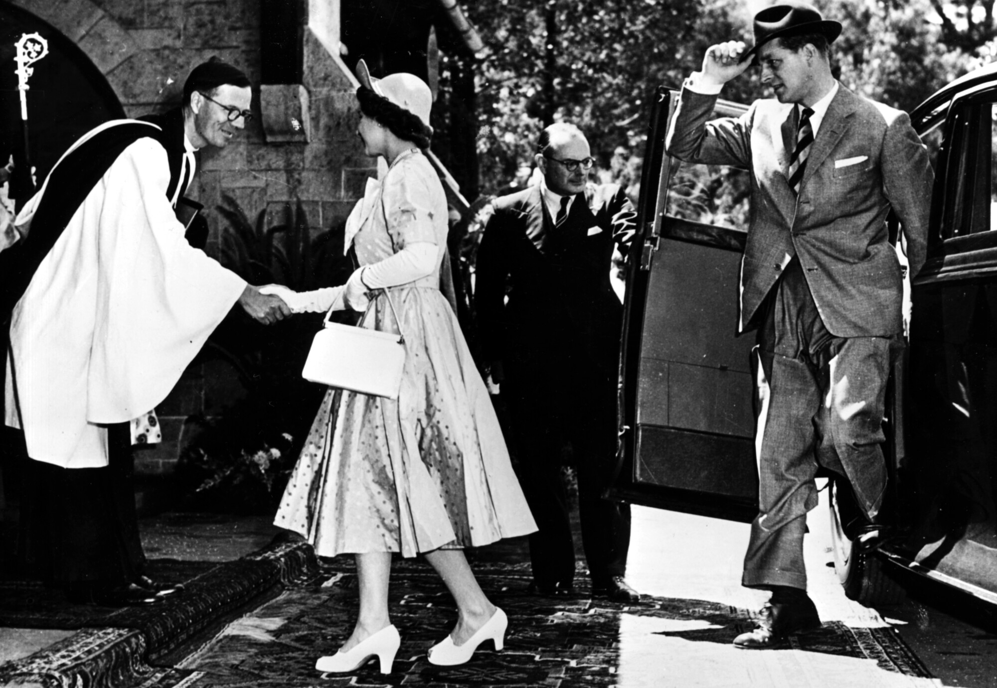 A man in clergy robes, left, shakes the hand of a woman in a dress and hat, as a man on the right emerges from a car 