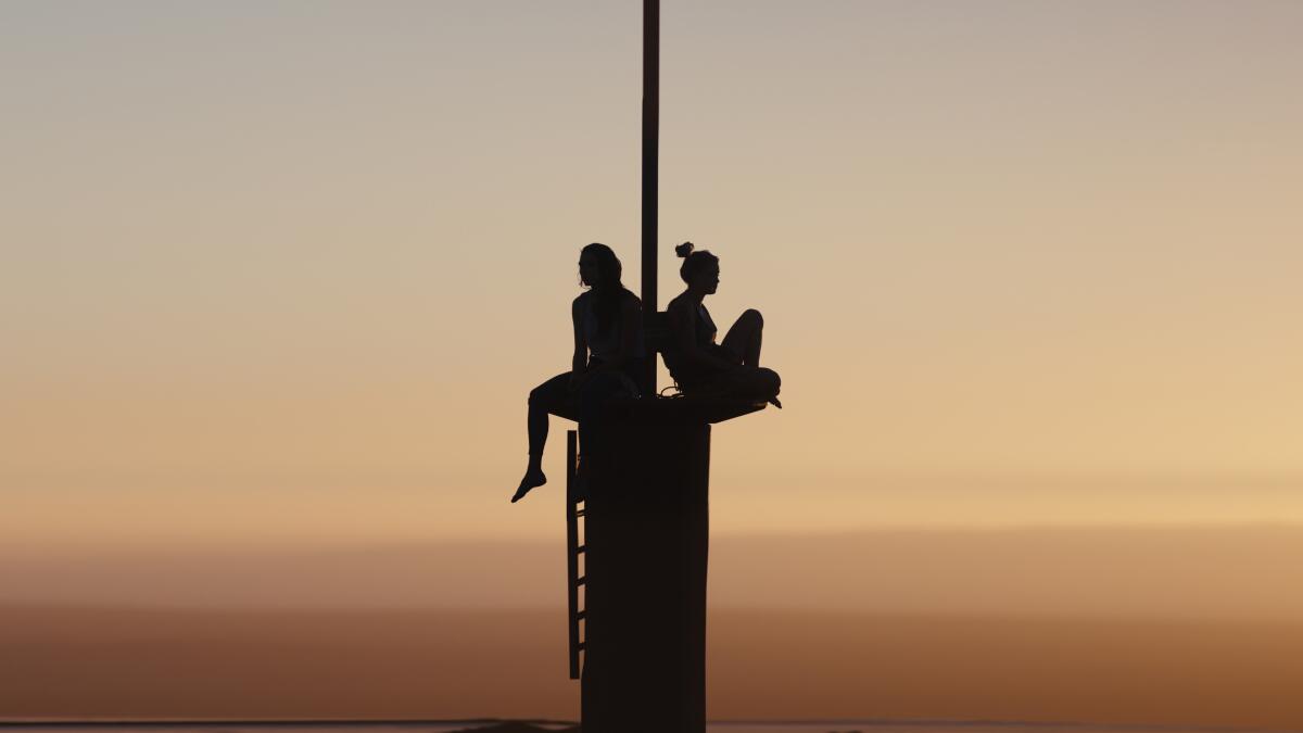 Two women sit on a tiny platform in the air as the sun sets.