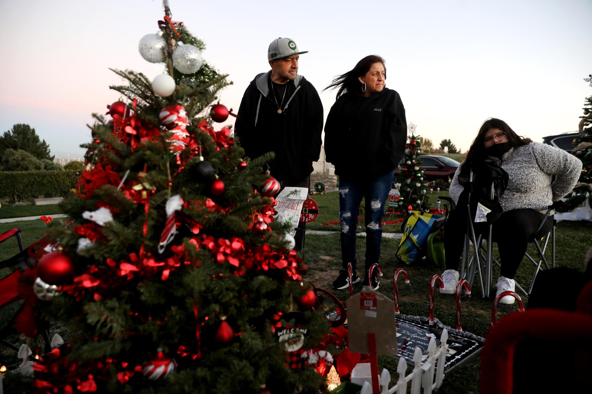 Three people, hands in jackets and hair blowing in the wind, gather near a decorated Christmas tree at a gravesite.