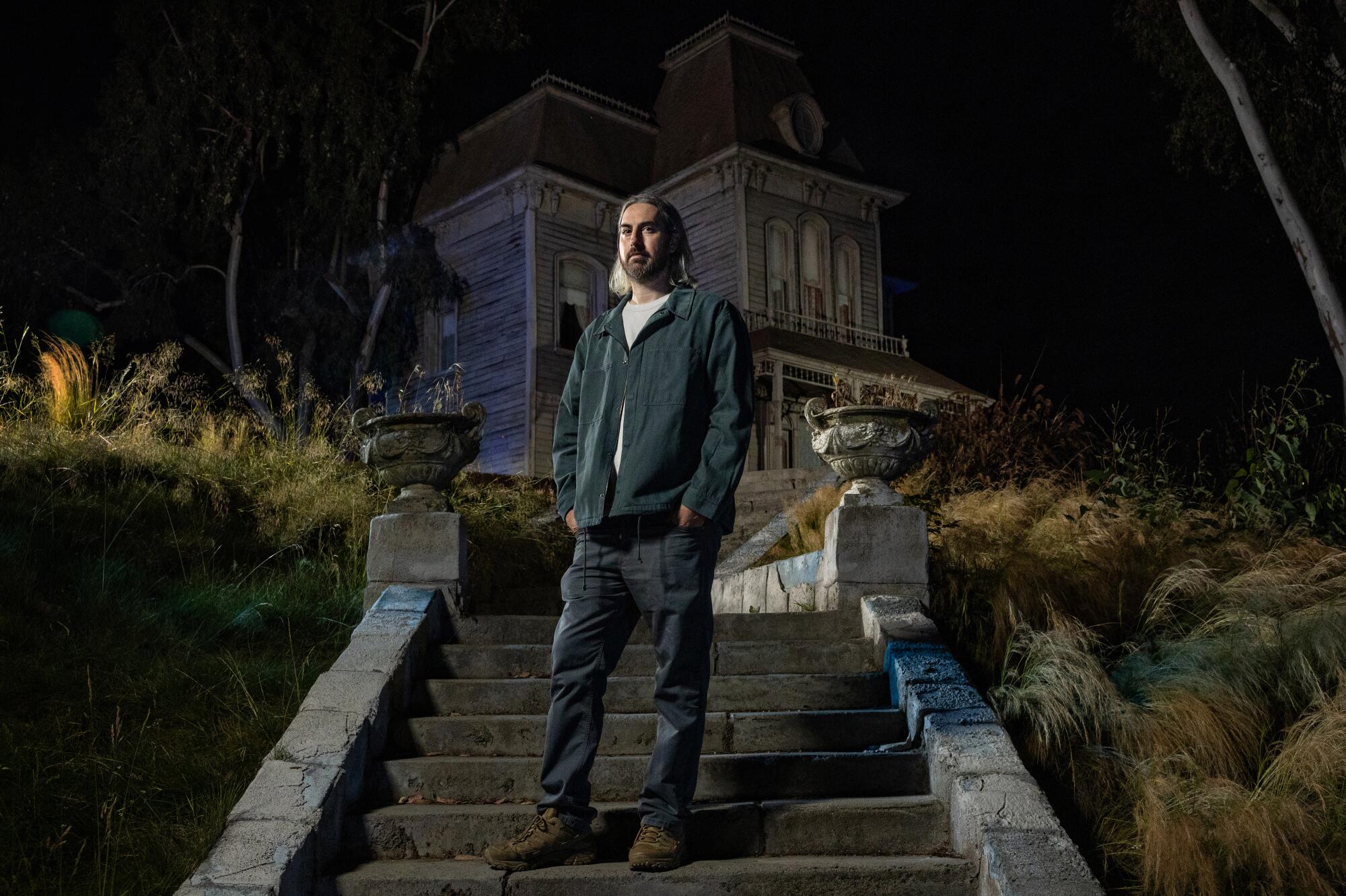 A man stands on the steps in front of a spooky gothic house at night.