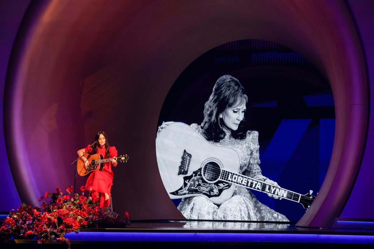 A woman in a red dress sitting and playing guitar in front of a mic and giant black-and-white image of a woman playing guitar