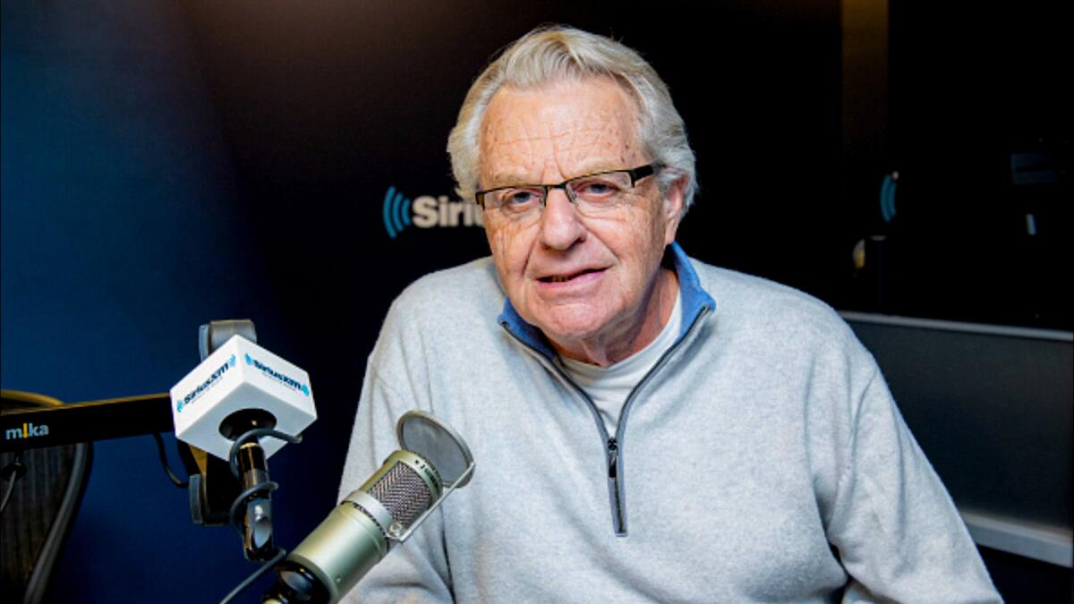 Jerry Springer sits behind several radio microphones wearing casual attire