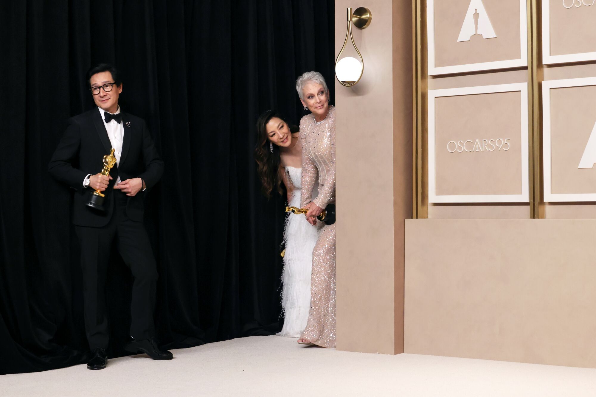 Three people in evening wear - a man stands at left while two women peek in from behind a wall with the writing "Oscars." 