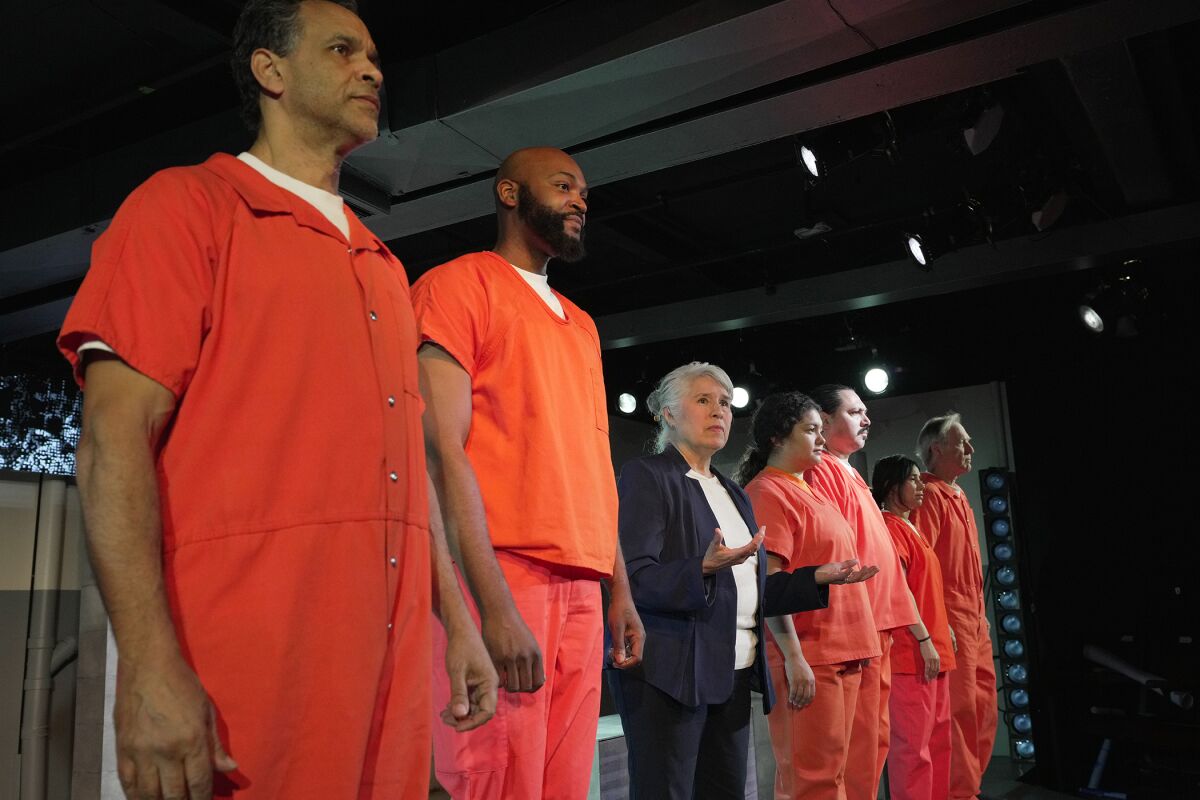 A woman speaks in the middle of six actors dressed in orange like prisoners.
