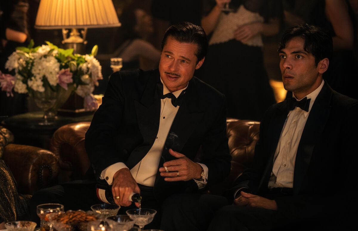 Two seated men wearing tuxedos. One is pouring champagne into a glass on the table before them. 