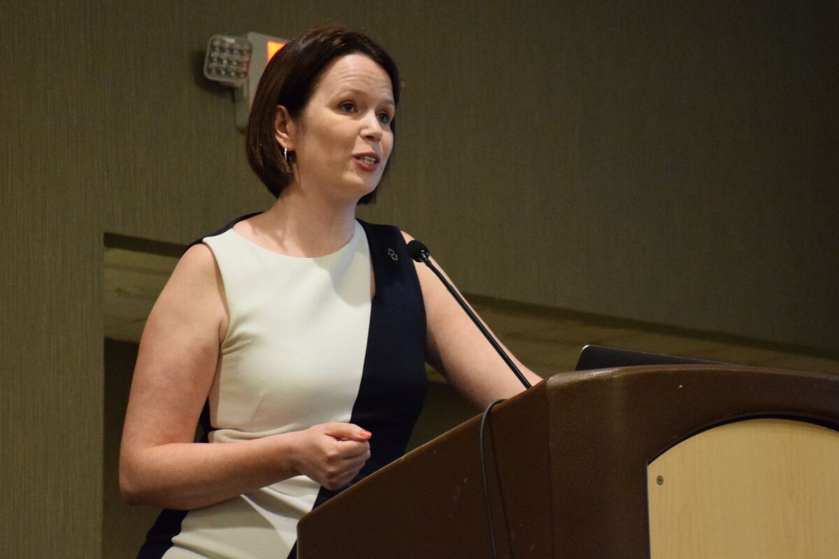 a woman in a cream and blue dress speaks behind a podium in a conference room