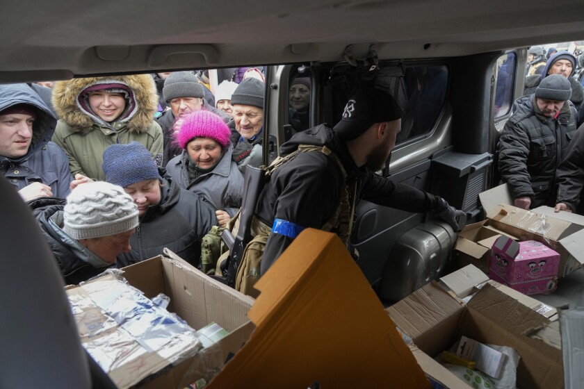 People in winter clothes and knitted hats gather in front of the open door of a car with open boxes inside 