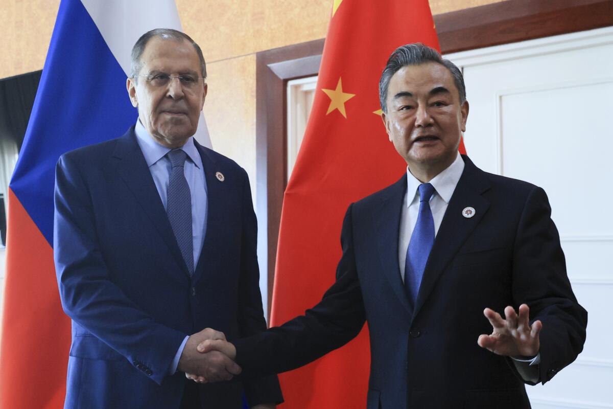 Two men in suits shake hands in front of flags 